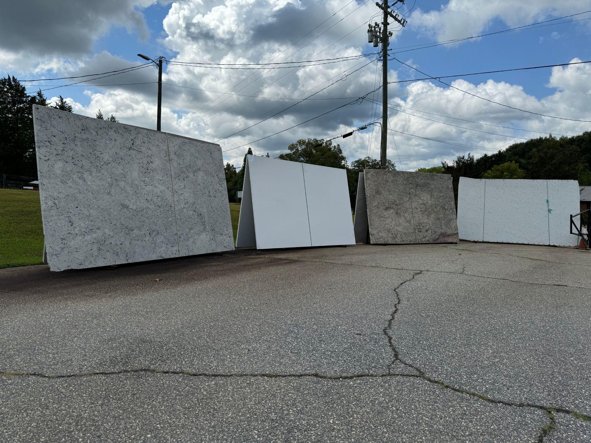 Natural stone slabs on display. The slabs feature varying shades of white and gray, with intricate patterns. A few cars can be seen in the background, showcasing the prominent East Coast Granite & Tile roadside location of the Travelers Rest showroom.