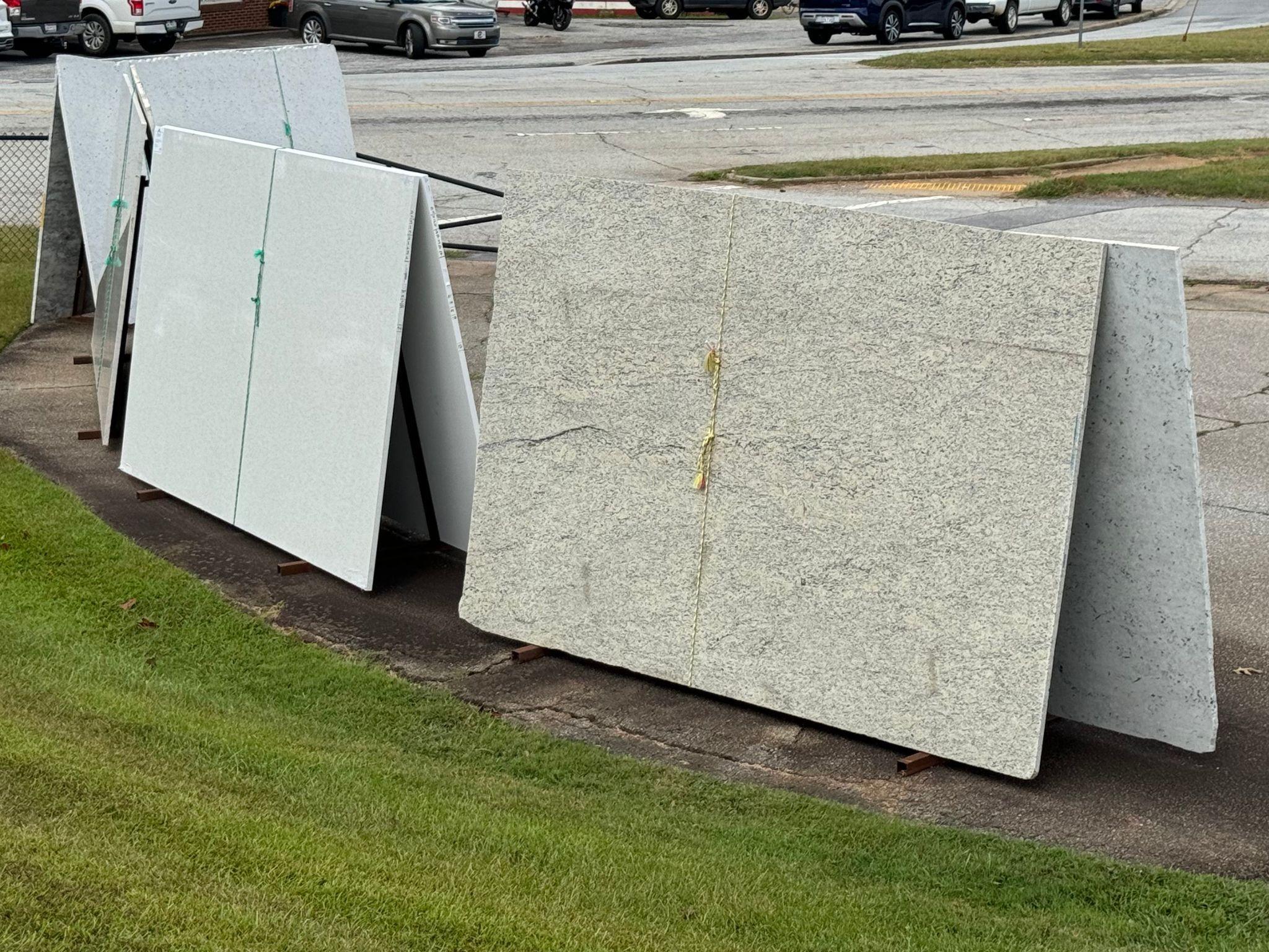 A further selection of slabs on display outside, including a mix of light and dark tones. Some slabs have solid colors, while others feature complex, veined patterns. The cloudy sky creates a soft, neutral backdrop for the display area.