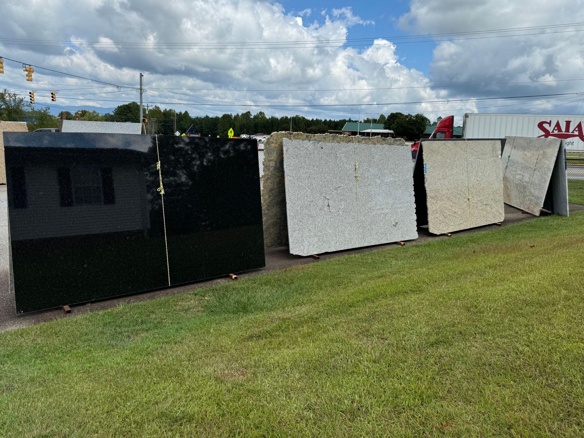 Large countertop slabs arranged in a row, including highly polished black slabs and lighter-toned stone with natural swirling patterns. The display is situated on the showroom’s paved lot, with patches of grass in the background.