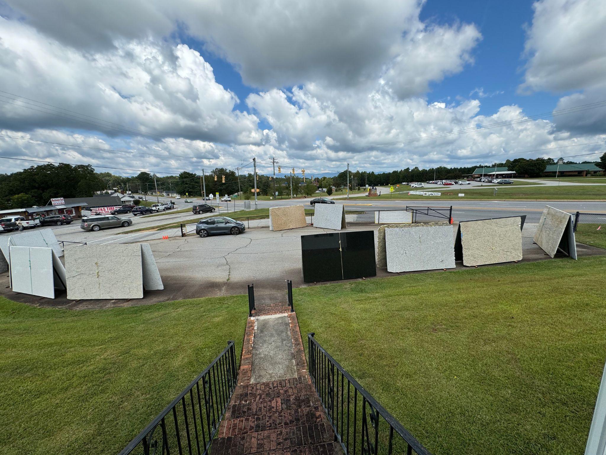 A wider-angle view of multiple large slabs displayed side-by-side in the parking area, showcasing various stone colors and patterns. The showroom building can be seen in the background, offering a sense of scale and location.